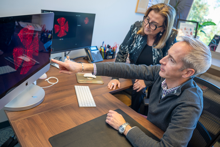 A professionally dressed man and woman sit behind a desk with computer monitors. The man points to a monitor that shows a retina on it.